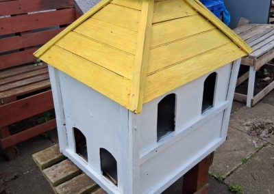 A homemade wooden aviary is painted white with a yellow roof
