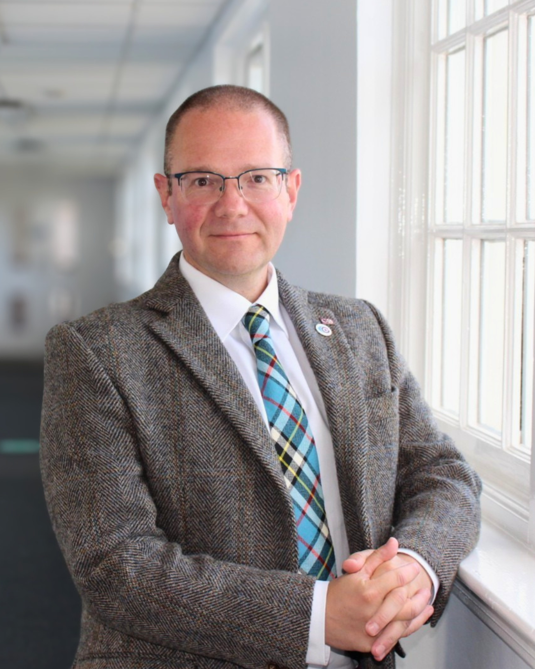 Andrew Thomson Carr Gomm A man (Andrew Thomson) wearing glasses and a tweed jacket stands in a hallway next to a window.