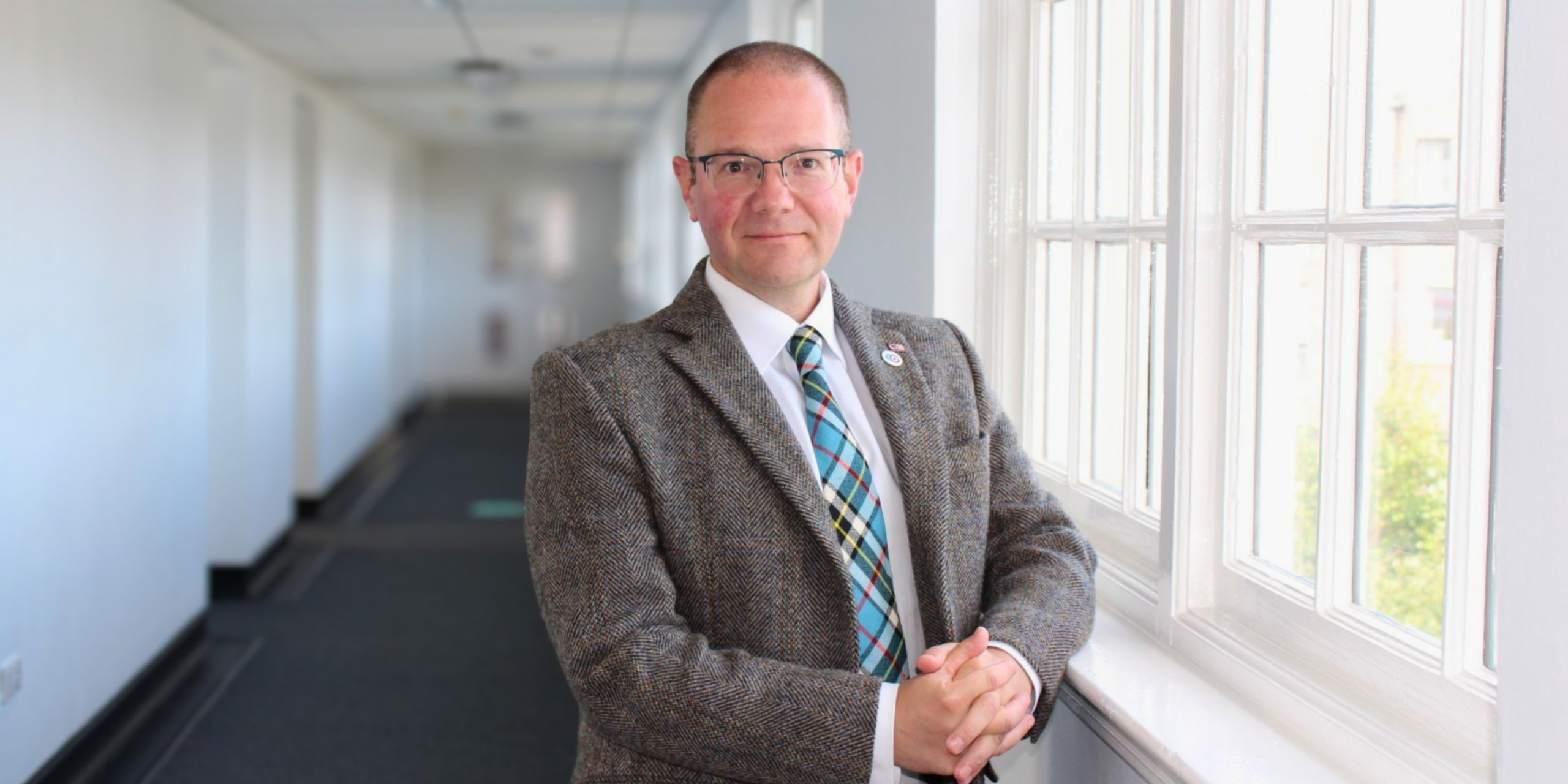 A man (Andrew Thomson) wearing glasses and a tweed jacket stands in a hallway next to a window.