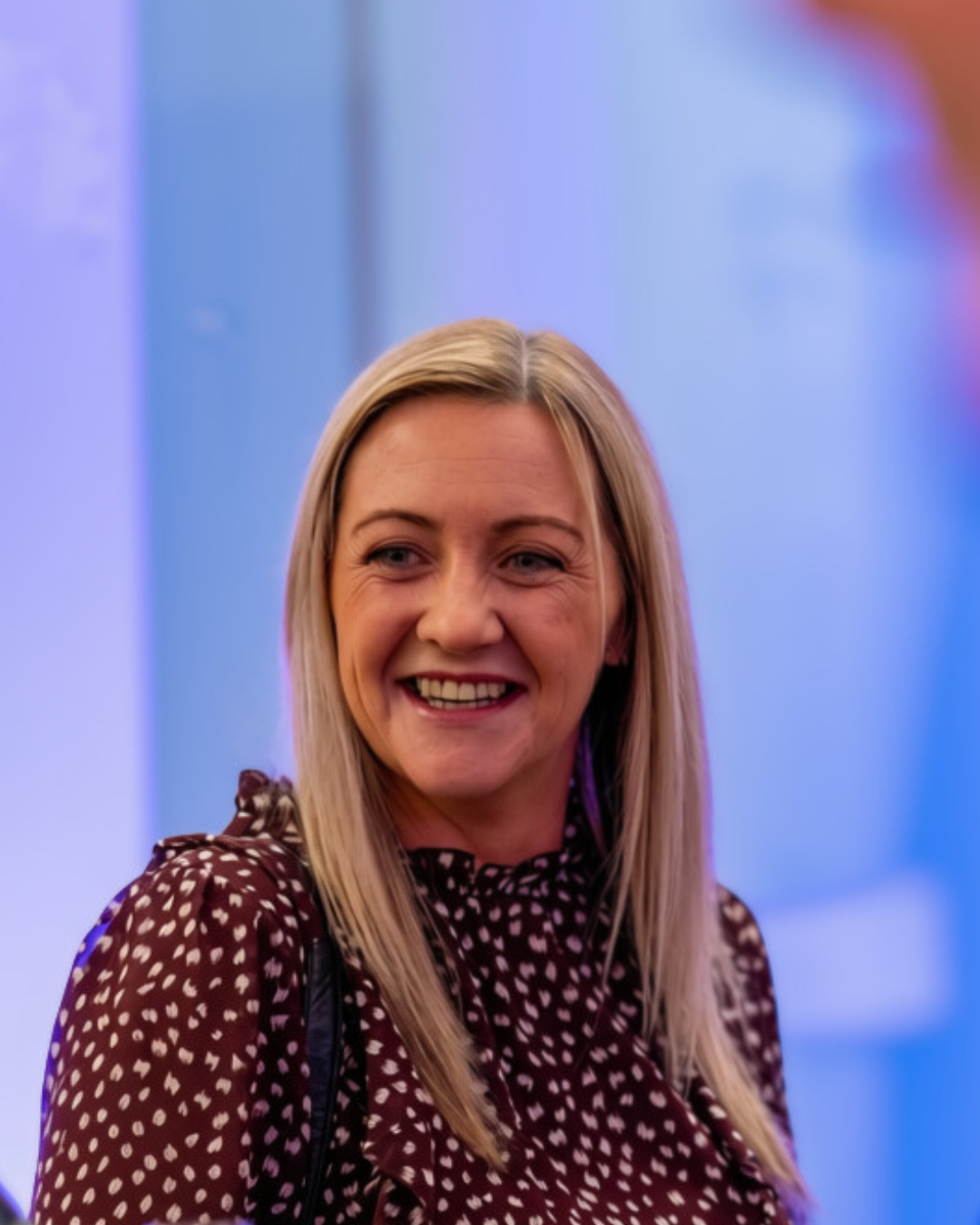 Fiona Lawrence Carr Gomm A woman (Fiona Lawrence) in a maroon dress with white polka dots smiles broadly/ She is in a softly lit room with a blue background.