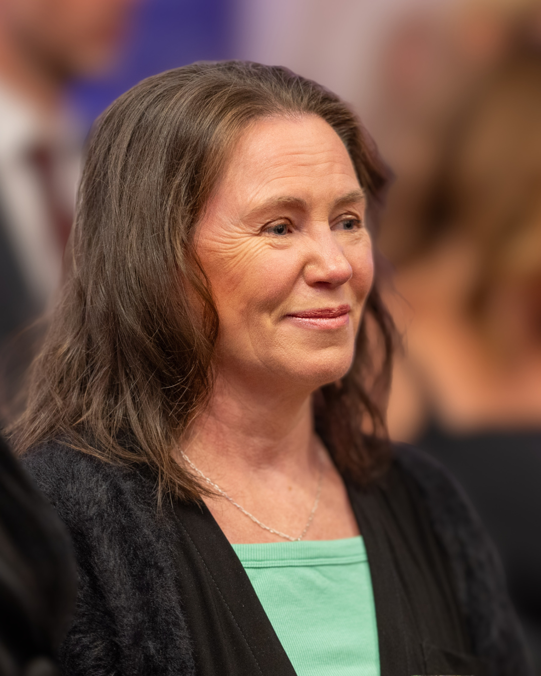 A woman (Leanne Wells) with wavy dark hair against a soft blurred background. She is wearing a light green top and black cardigan
