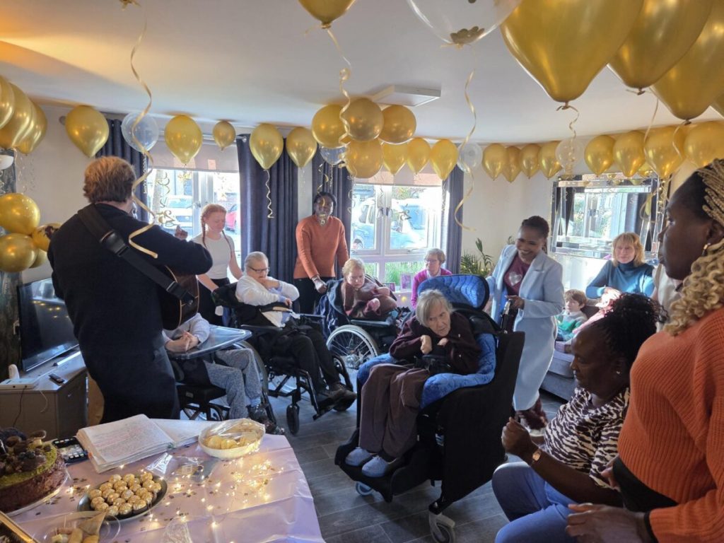 A vibrant party scene with a room decorated with gold balloons. There is a musician playing the guitar while women who are wheelchair users watch on with their support staff | Working in social care
