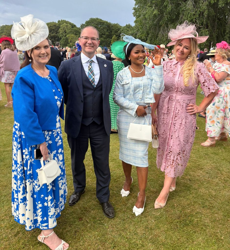 A group of four people stands outdoors on grass, dressed in formal attire and various hats. The woman on the left (Asiya) wears a blue outfit with a floral pattern, while the woman on the right (Ruth) is in pink lace. The individuals in the middle (Andrew & Busayo) are dressed in a suit and a light blue outfit. A garden scene and other people are visible in the background.