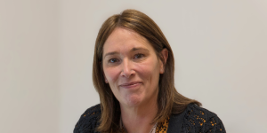 A woman (Susan Colin) with brown shoulder length hair against a neutral wall