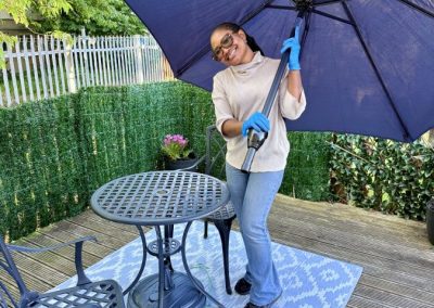 An African woman, Busayo, holds up a large sun umbrella. She's stands by garden furniture on decking.