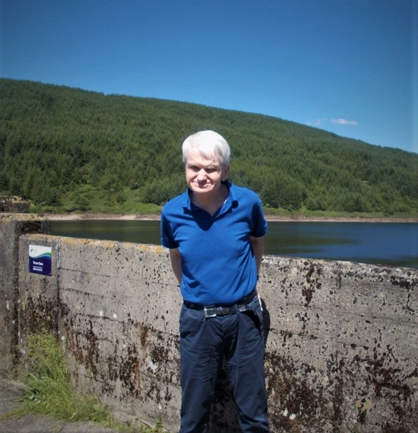 A man (James) wearing a blue tshirt and jeans stands next to a reservoir. There is a loch and hills in the background.