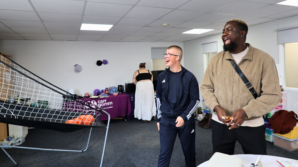 A room with two people smiling and standing near a basketball game setup. A table with a purple Carr Gomm banner is at the back. The space has white walls and a gray carpet.