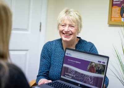 A woman smiles warmly while holding up a laptop in an office environment. | Carr Gomm HR Officer