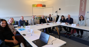 A group of Trustees and Carr Gomm staff seated around a conference table in a bright meeting room, with name placards and water bottles, one laptop open in the foreground.