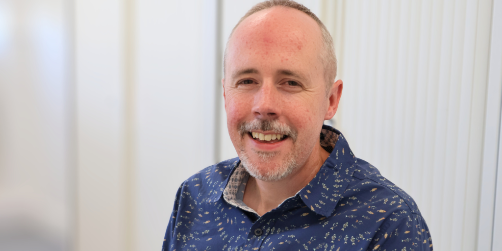A man (Paul Marshall) with a beard sits in an office environment wearing a navy shirt.