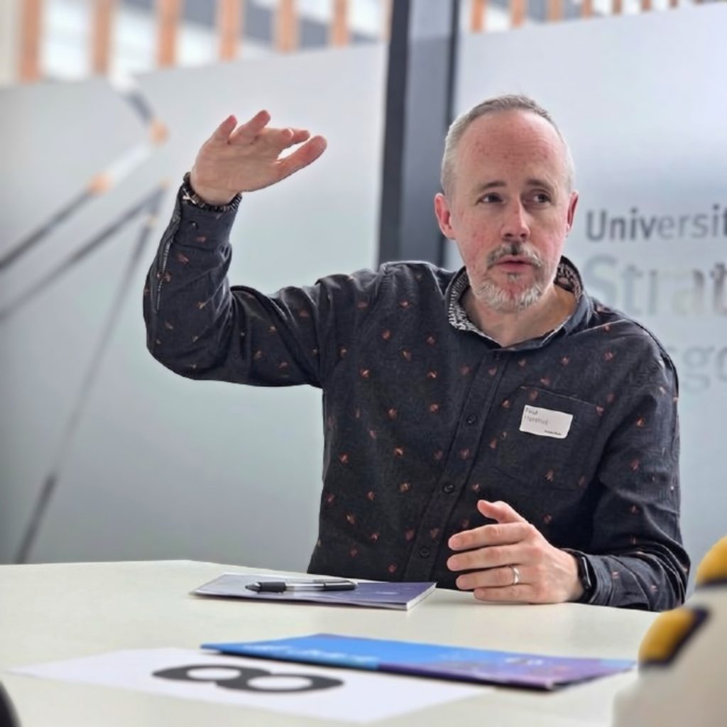 A man (Paul Marshall, Deputy Chief Executive) in a dark patterned shirt gestures with his hands while speaking at a table with blue and white papers; a university sign is visible in the background.