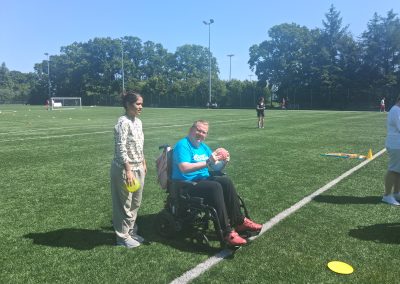 A woman sits in a wheelchair on a football pitch holding a ball. A woman stands beside her.