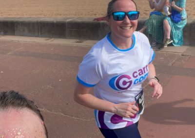 Two women running on a promenade by the sea. They are both wearing sunglasses, one is wearing a Carr Gomm tshirt.
