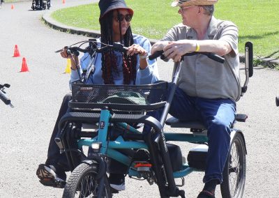 Two people cycling an adaptive bicycle on a tarmac track. There are cones on the track in the background.