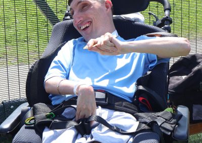 A man (Cameron) wearing a t-shirt and shorts sits in a wheelchair by a sportsfield.