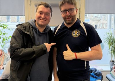A man wearing a Scotland football jersey stands next to another man in an office environment. They are both smiling with thumbs up.
