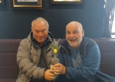 Two men smile while holding a vase with flowers.