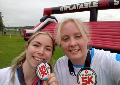 Two women smiling and holding up medals. In the background is an inflatable obstacle course.