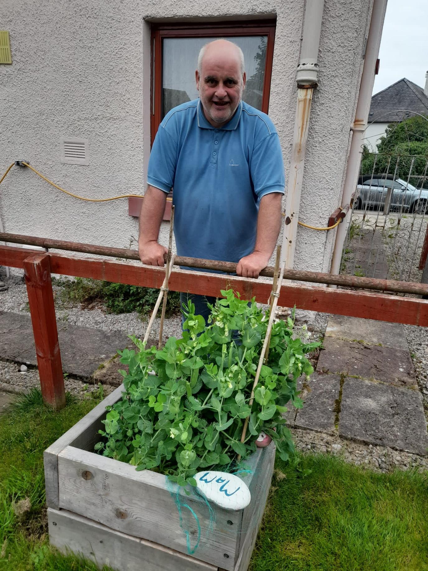 A man (Anthony) wearing a blue t-shirt stands proudly behind a raised planter. The planter has a variety of vegetables.
