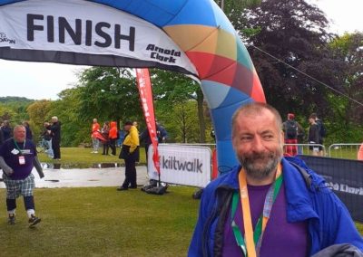 A man (Robert) wearing a Kiltwalk lanyard stands by the Kiltwalk finish line.