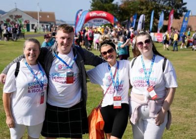 A group of 4 people hugging each other. They are wearing Carr Gomm tshirts and in the background are people and the Kiltwalk finish line.