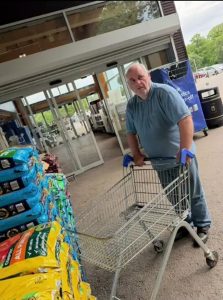 A man (Ant) wearing a blue tshirt and grey joggers stands by sacks of compost at the entrace to a store. He is pushing a trolley.