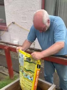 A man (Ant) tipping a yellow bag of compost into a raised planter bed.