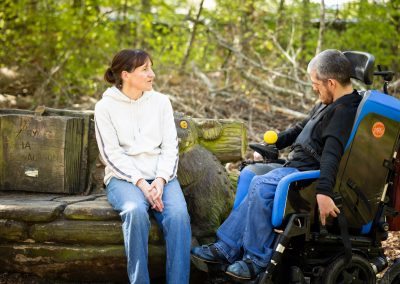 A female Support Practitioner sits outside on a bench next to a man who is a wheelchair user.