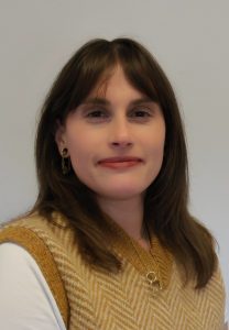 A woman (Ekaterina) with shoulder length brown hair wearing a mustard knitted vest and white tshirt.