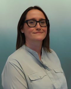 A woman (Gillian Lewis) with dark hair and glasses wearing a light blue button-up shirt, standing against a plain white background with a neutral, slight smile.