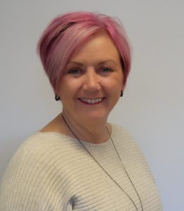 A woman (Mandy Hay) with pink highlights wearing a diamond necklace against a soft blurred background.