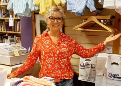 A woman (Ania) with glasses wearing a red shirt smiles in front of lots of hand knitted baby cardigans. She is in the Carr Gomm shop in Inverness. Carr Gomm Shop.