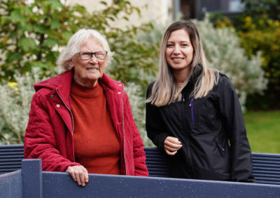 Two women stand out side with greenery in the background. The lady on the left is older and wearing red. The woman on the right is younger and wearing black. | Carr Gomm Seaboard Care at Home