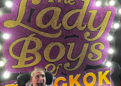 A woman in a wheelchair is smiling in front of a large sign reading 'The Lady Boys of Bangkok'