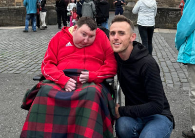 A man in a wheelchair is next to a man kneeling. They are in front of Edinburgh Castle.