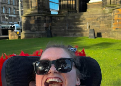 A women in a wheelchair smiles at the camera. She is in front of the Scotts Monument.